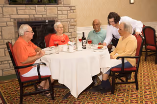 Four elderly people sitting around a round dining table covered with a white tablecloth in a room with a stone fireplace. A waitress is serving one of the elderly individuals. The table has a bottle of wine, glasses, and plates with food.