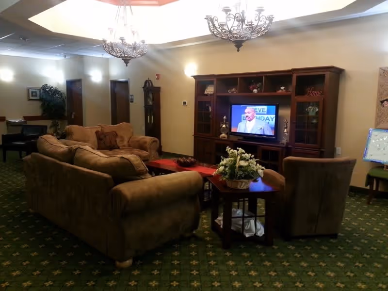 A cozy living room area with brown upholstered sofas and chairs arranged around a wooden coffee table and side table with a flower arrangement. A large wooden entertainment center holds a flat-screen TV displaying a man speaking. The room has green carpet with a patterned design, beige walls, and soft lighting from wall sconces and ceiling fixtures. There is a grandfather clock and some framed pictures on the walls.