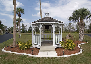 White wooden gazebo with a shingled roof situated on a grassy area surrounded by palm trees and small bushes, with a paved pathway leading to it under a partly cloudy sky.