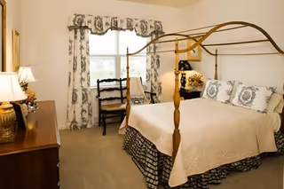 A bedroom with a four-poster bed covered in a beige quilt and decorative pillows. There is a wooden dresser with a lamp on it to the left, and two wooden chairs near a window with patterned curtains. The room has beige carpeting and light-colored walls.