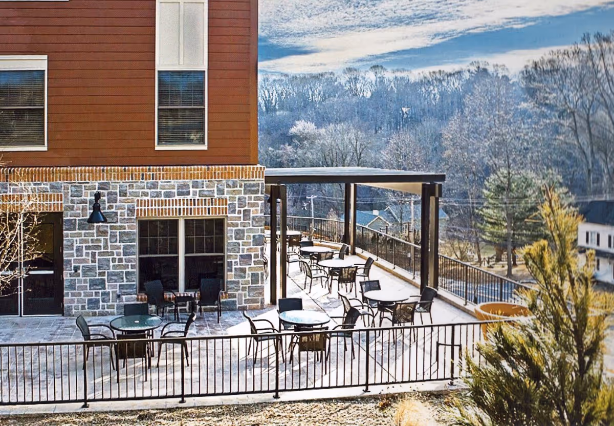 Outdoor patio area at Plush Mills Senior Living with multiple round tables and chairs arranged under a pergola and open sky. The building exterior features a combination of stone and wood siding. Trees and houses are visible in the background under a partly cloudy sky.