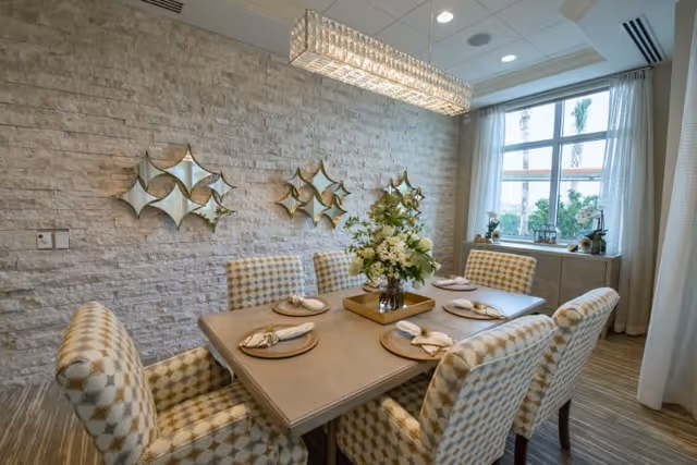 A dining room with a rectangular table set for six people with beige and white patterned chairs. The table has a centerpiece with a floral arrangement on a gold tray. The room features a textured stone accent wall with decorative mirrors and a large window with white curtains letting in natural light.