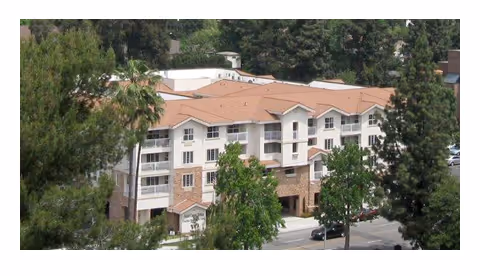 Three-story apartment-style senior living building with balconies and a red-tiled roof surrounded by trees.
