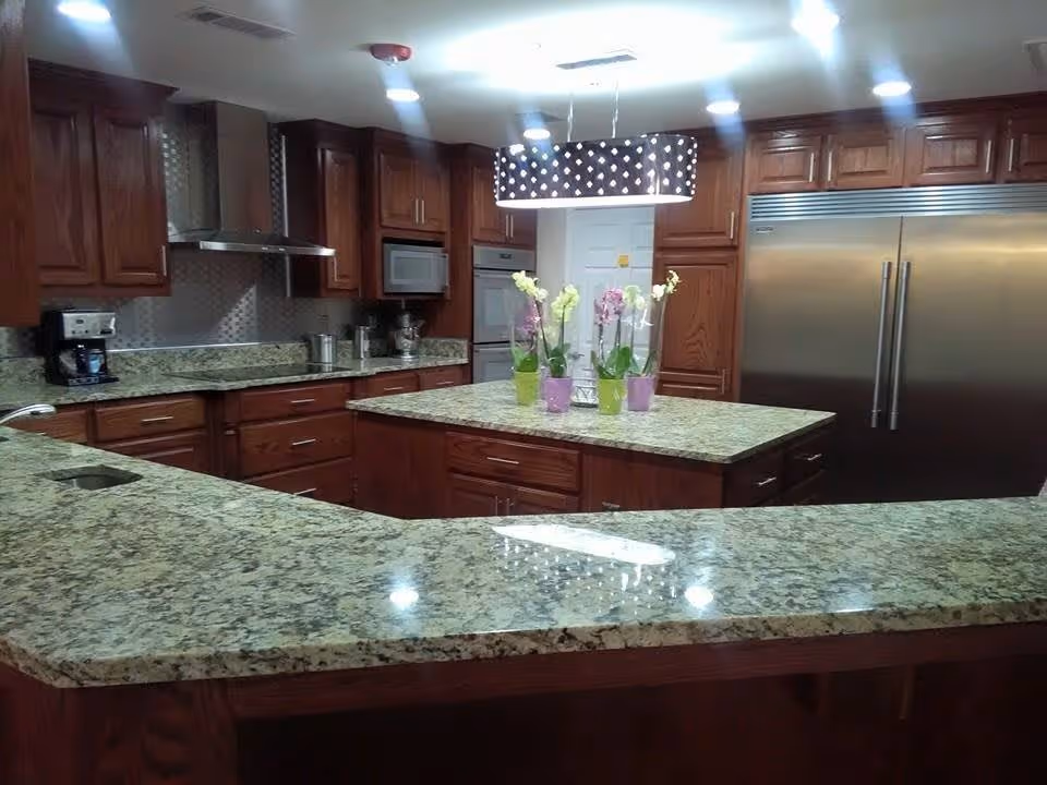A spacious kitchen with granite countertops and wooden cabinets. The kitchen features a large island in the center with several potted orchids on top. Stainless steel appliances including a double-door refrigerator, built-in oven, microwave, and a coffee maker are visible. The ceiling has recessed lighting and a decorative hanging light fixture above the island.
