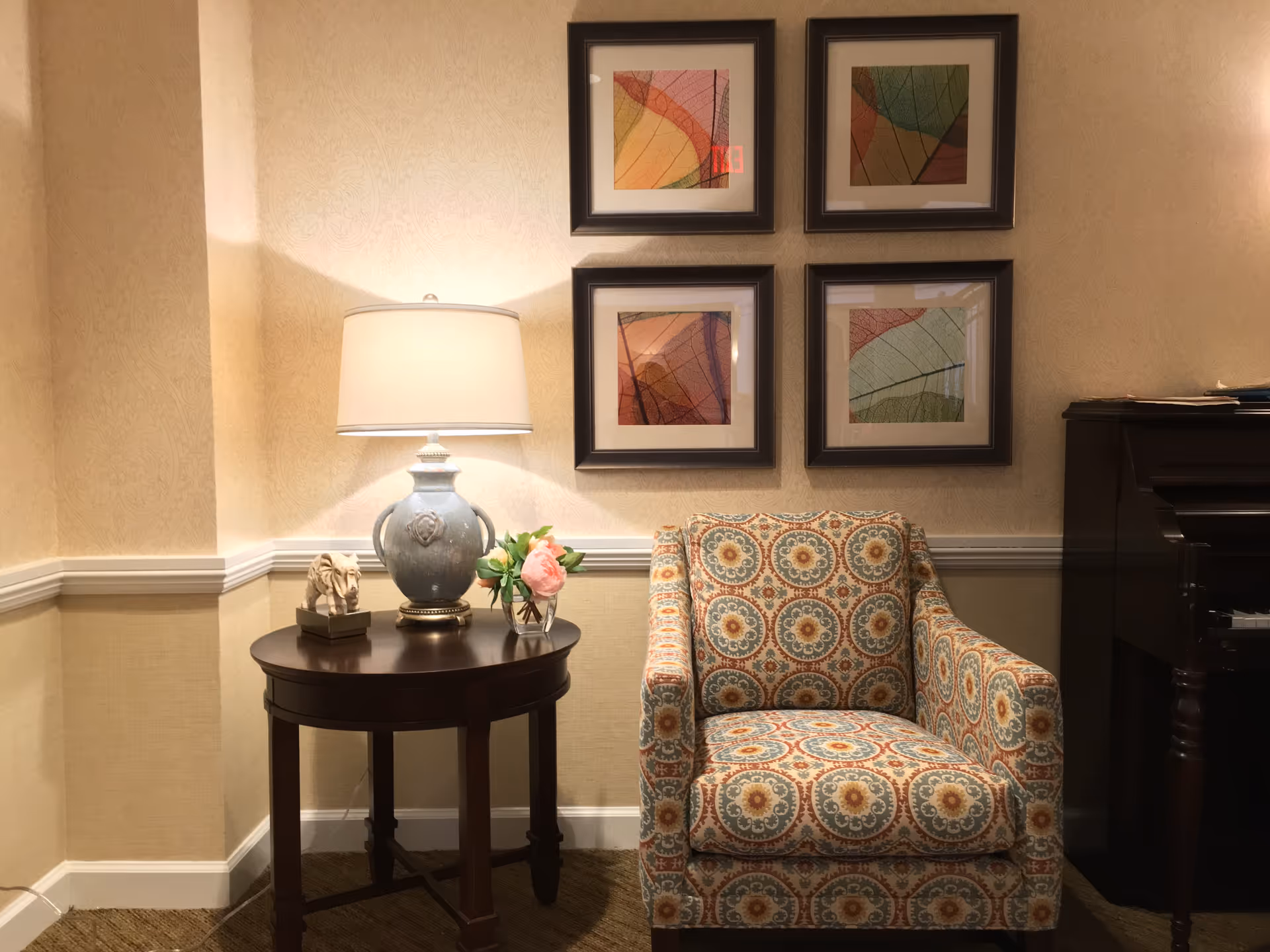 A cozy corner with a patterned armchair next to a dark wooden side table holding a decorative lamp, a small horse sculpture, and a glass vase with pink flowers. Four framed abstract leaf prints hang on the beige patterned wall above the chair and table. Part of a dark wooden piano is visible on the right side.