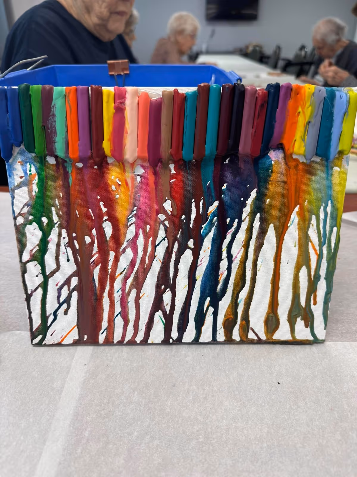 A colorful melted crayon art piece on a canvas with crayons glued at the top, melting downwards creating vibrant streaks of color. In the background, elderly people are seated and engaged in activities in a communal room.
