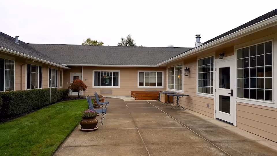 Outdoor courtyard area of a senior living facility with beige siding buildings surrounding a concrete patio. There are several metal chairs with cushions along the left side near a green hedge, a potted plant, and a small tree. The patio has a table and bench along the right side near large windows and a white door.