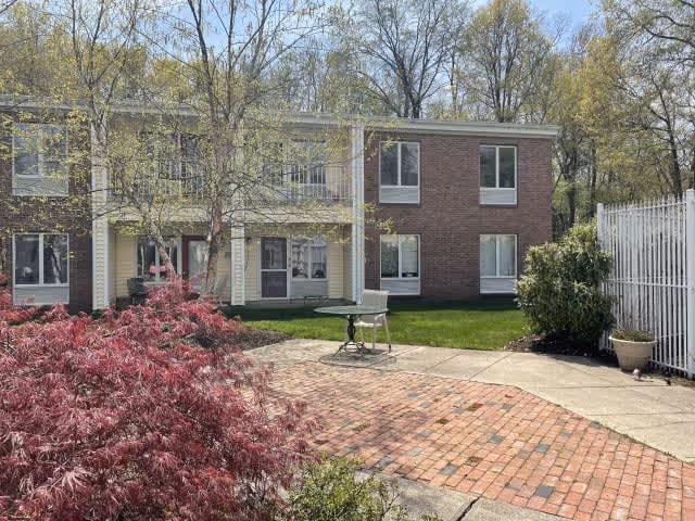 Outdoor view of a senior living facility showing a two-story building with brick and siding exterior, surrounded by trees and greenery. There is a paved patio area with a small table and chair, and a white fence on the right side.