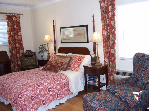 Bedroom with a bed dressed in red patterned bedding, two bedside tables with lamps, a patterned armchair and floral curtains.