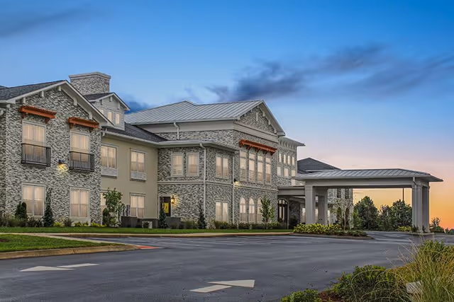 Exterior view of The Blake at Kingsport senior living facility during sunset, showing a large two-story building with stone and stucco facade, multiple windows, a covered entrance, landscaped greenery, and a paved driveway with directional arrows.
