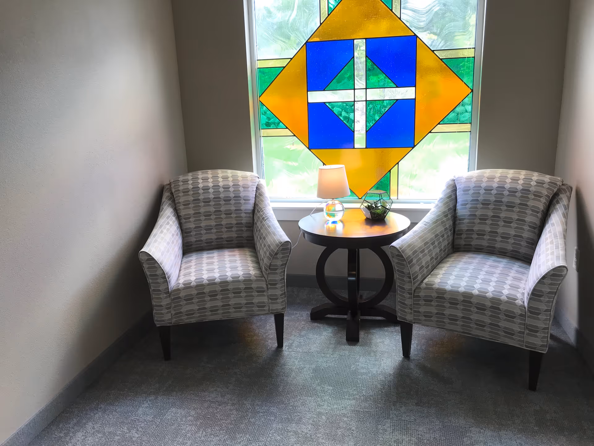 Two patterned armchairs positioned on either side of a small round wooden table with a lamp and a small plant, set in front of a window featuring a colorful stained glass design with blue, yellow, and green geometric shapes.