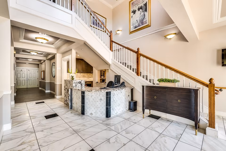 Bright and spacious reception area in a senior living facility with a marble-tiled floor, a curved reception desk under a staircase with wooden handrails, a dark wooden cabinet with a plant on top, and framed artwork on the wall.