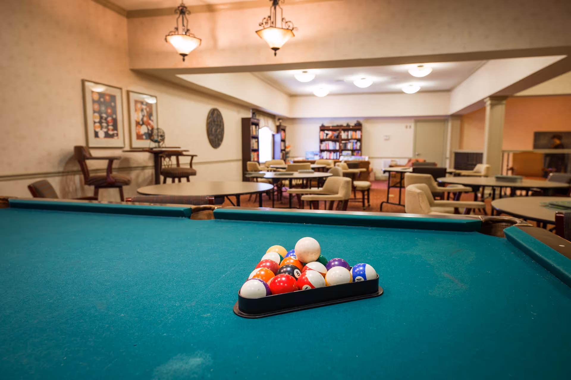 A recreational room with a pool table in the foreground, set up with billiard balls in a triangle rack. In the background, there are several round tables with chairs, bookshelves filled with books, and framed artwork on the walls. The room is warmly lit with ceiling lights and hanging lamps.