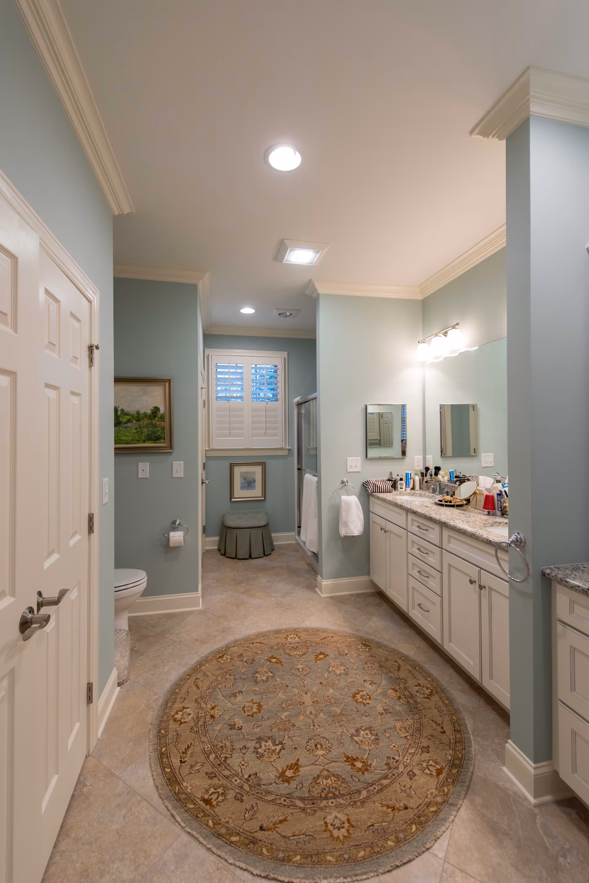 A spacious bathroom with light blue walls, a large round patterned rug on the tiled floor, a double sink vanity with granite countertop and various toiletries, a toilet, a small cushioned stool under a window with white shutters, and a glass-enclosed shower in the background.