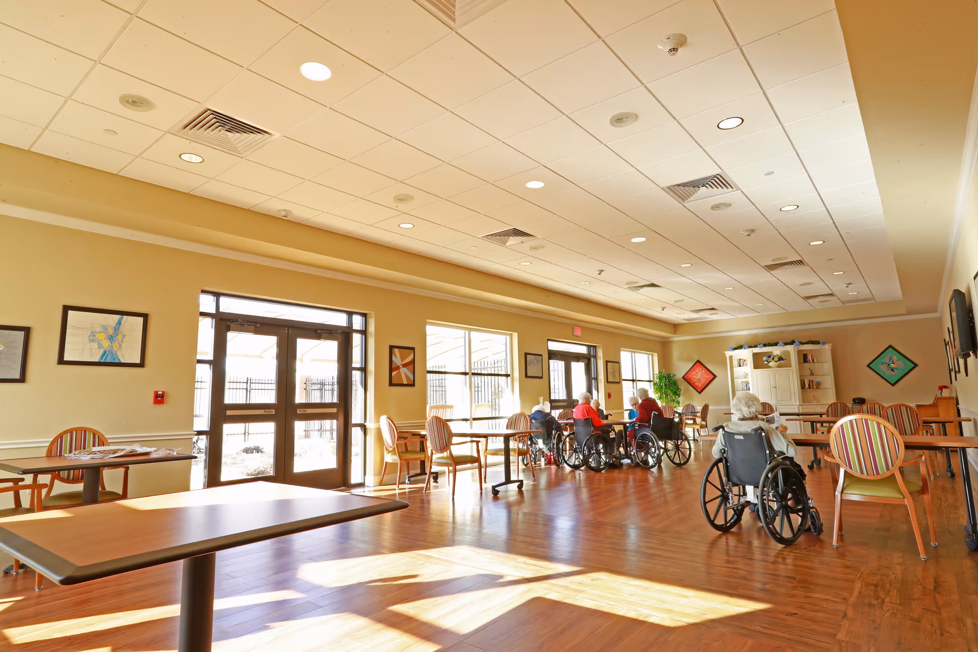 Sunlit communal dining/activity room with tables, chairs, and several elderly residents in wheelchairs near the windows.