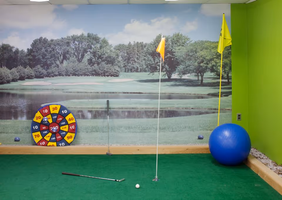 Indoor putting green area with a golf ball, two golf clubs, two flags with holes, a colorful dartboard on the wall, and a large blue exercise ball. The wall has a mural of an outdoor golf course with trees and a pond. The room has bright green walls and a green carpet floor.