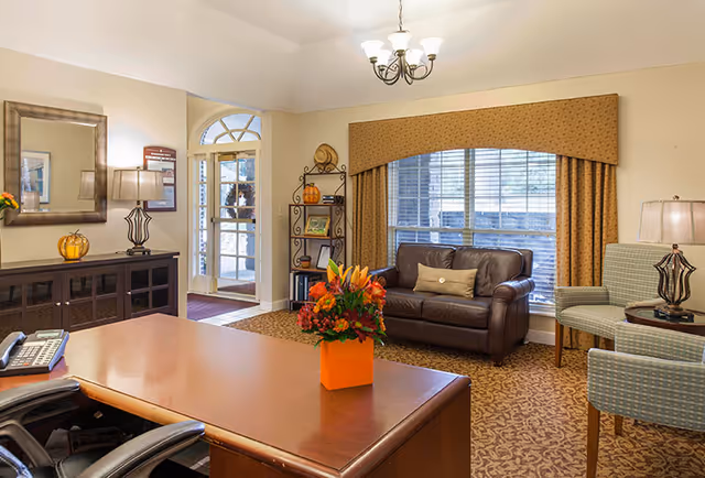 A well-lit office or reception area with a wooden desk in the foreground, a bouquet of flowers in an orange vase on the desk, a leather couch with a pillow, two upholstered chairs, a window with blinds and patterned valance, a side table with a lamp, a decorative shelving unit with framed photos and pumpkins, and a door with glass panels leading outside.