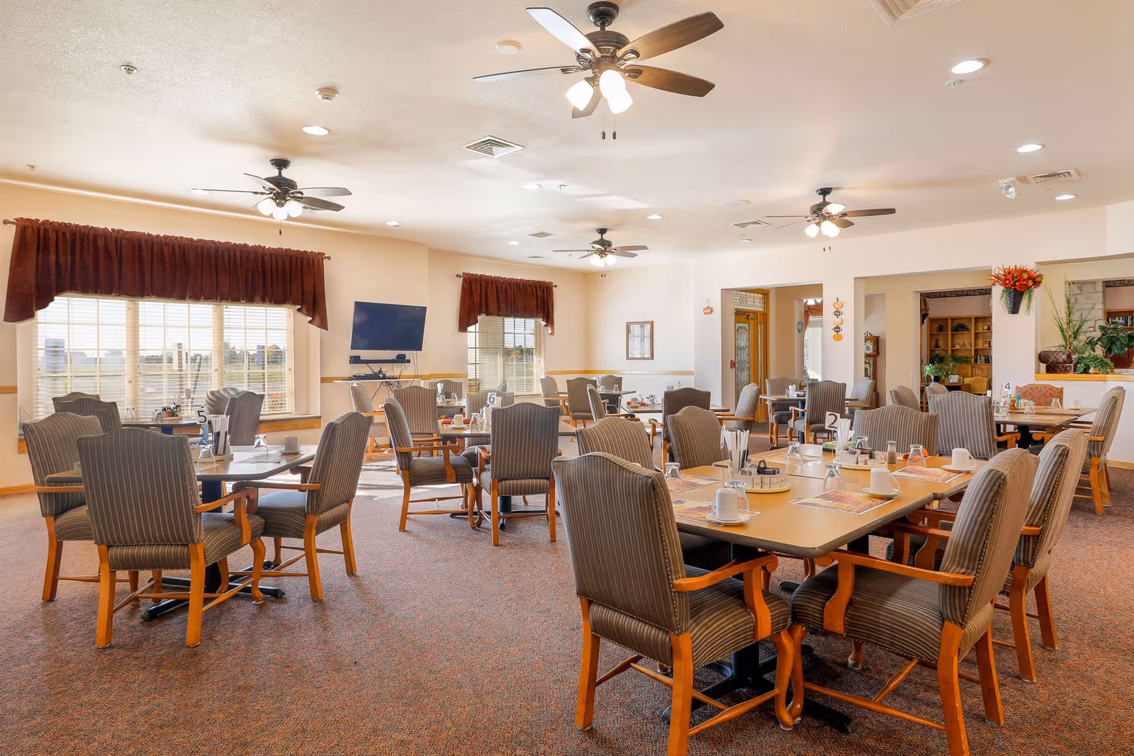 A spacious dining room with multiple tables and cushioned chairs arranged neatly. The tables are set with cups, plates, and condiments. Large windows with maroon valances allow natural light to fill the room. Ceiling fans with lights hang from the ceiling, and a flat-screen TV is mounted on one wall.