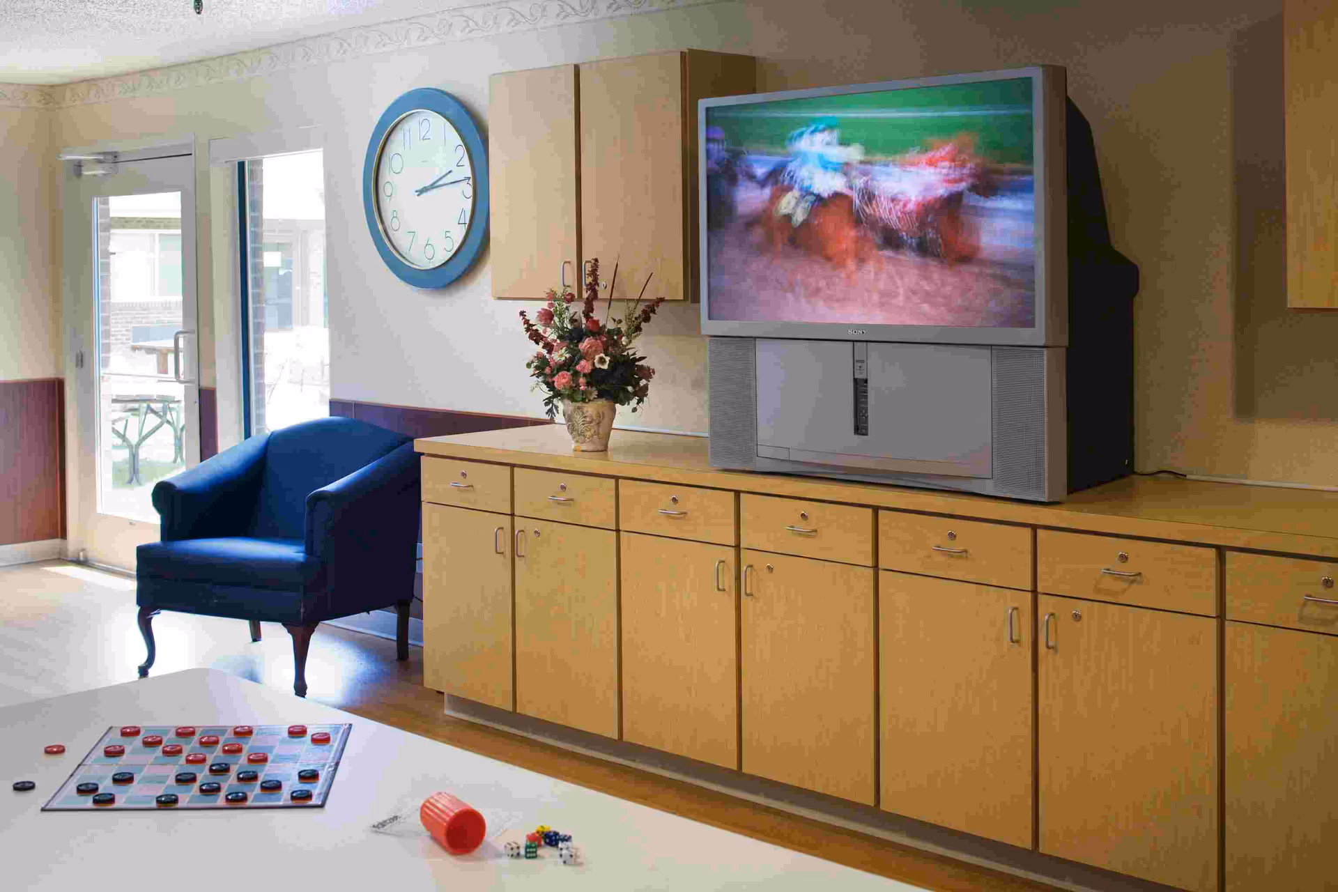 A cozy living room area in the Logan Center featuring a large wooden cabinet with multiple drawers and a vintage Sony television on top showing a blurred horse race. A blue armchair is placed near a glass door leading outside. A round wall clock with a blue frame hangs above the cabinet, and a vase with flowers is also on the cabinet. In the foreground, a table holds a checkers board with pieces and some dice scattered around.