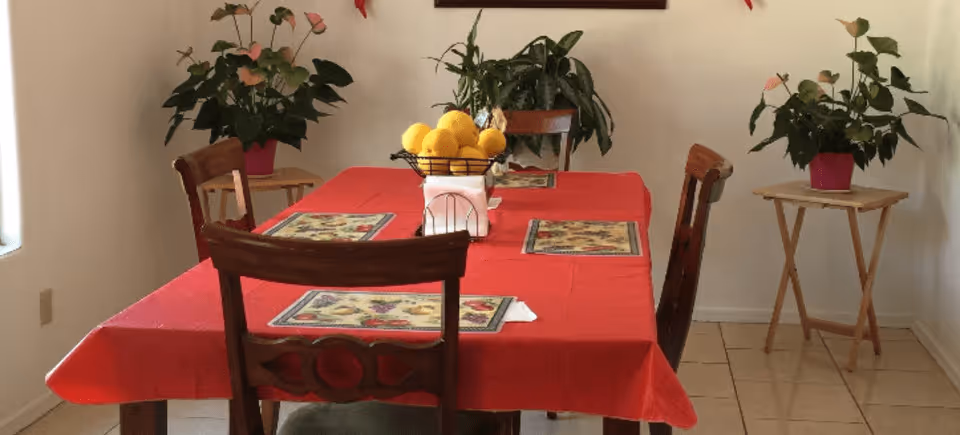 A dining room with a rectangular table covered with a red tablecloth. Four wooden chairs surround the table, which has four placemats and a centerpiece bowl filled with yellow lemons. There are two small wooden side tables against the walls, each holding a potted plant with green leaves and pink flowers. The room has tiled flooring and white walls.