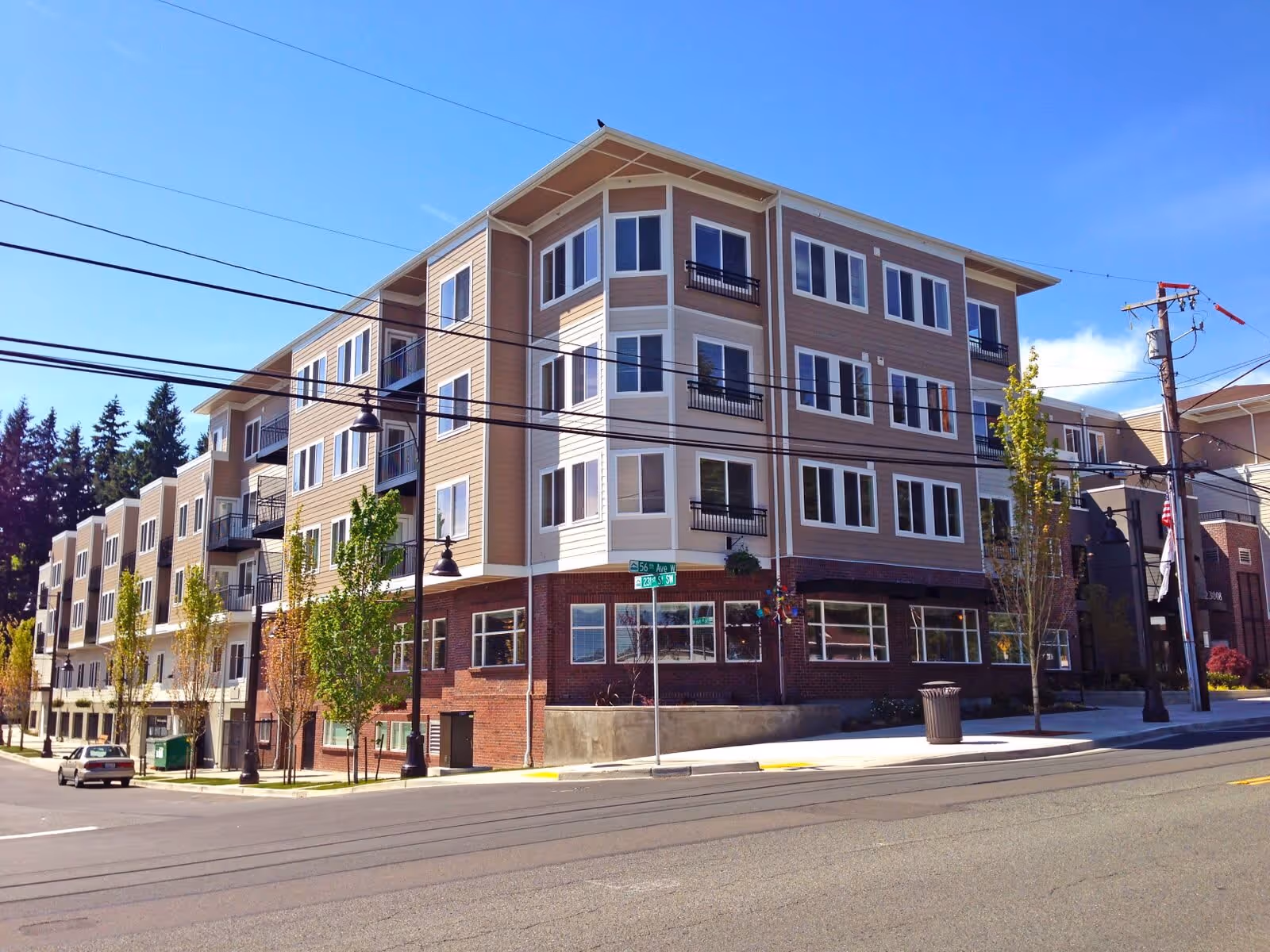 Exterior view of a multi-story residential building on a sunny day with clear blue sky. The building has beige and white siding with brick on the lower level and multiple windows. There are small trees planted along the sidewalk and a street intersection with visible street signs.