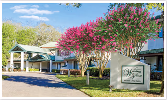 Exterior view of Weinberg Village Assisted Living facility with a driveway, green-roofed entrance, blooming pink flowering trees, and a sign displaying the facility name.