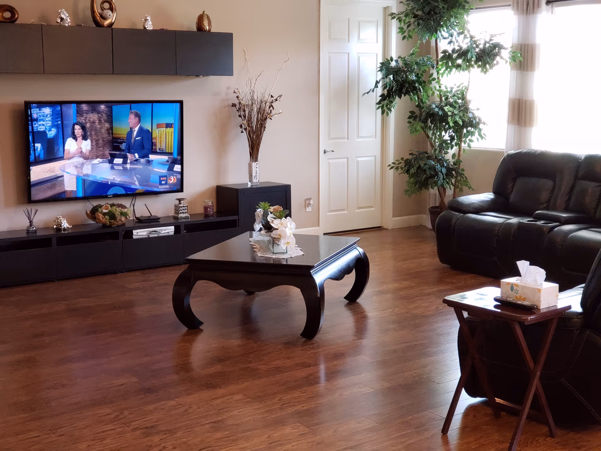 A cozy living room with wooden flooring, a black leather sofa, a black coffee table with decorative items, a side table with a tissue box, a large flat-screen TV mounted on the wall showing a news broadcast, a black entertainment unit with decorative items, a tall plant near a window with sheer curtains, and a closed white door.