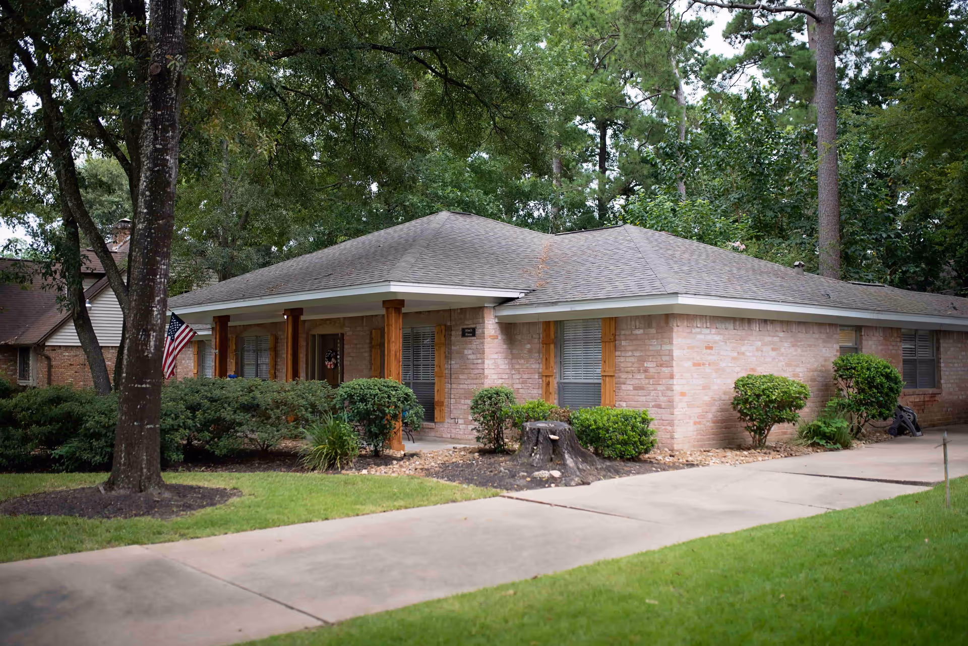 Single-story brick cottage with a covered front porch, wooden columns, shrubs, and an American flag by the driveway.