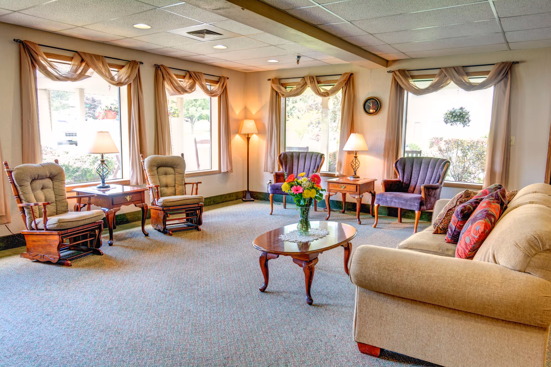 A bright and cozy living room in a retirement residence featuring a beige sofa with colorful cushions, two purple armchairs, two wooden rocking chairs with cushions, a wooden coffee table with a vase of flowers, and three large windows with sheer curtains letting in natural light. There are also two table lamps and one floor lamp providing warm lighting.