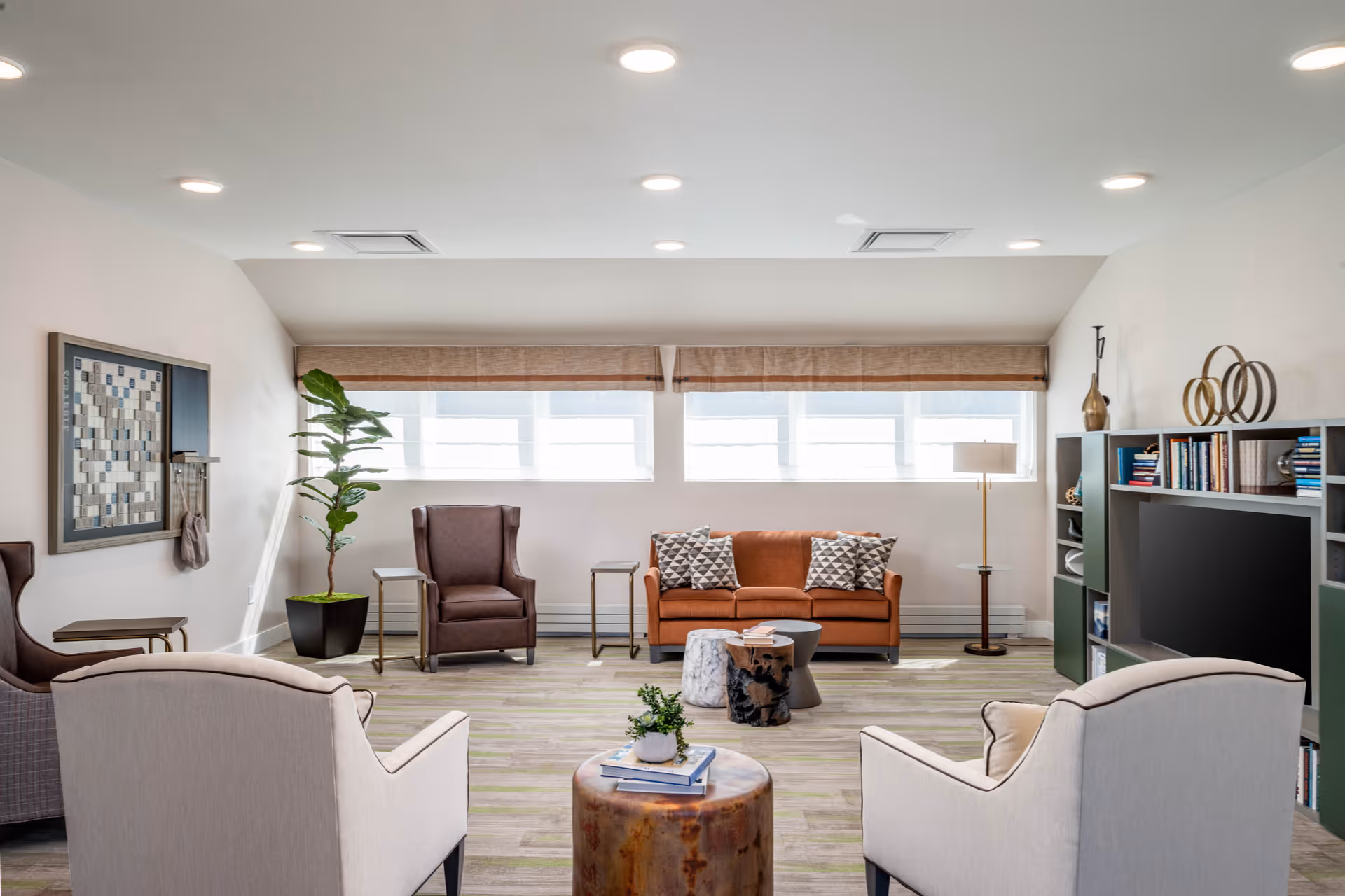 A bright and modern living room with a brown sofa, two beige armchairs, a brown armchair, a small round wooden coffee table with books and a plant, a floor lamp, a large TV on a bookshelf filled with books and decorative items, and large windows with blinds letting in natural light.