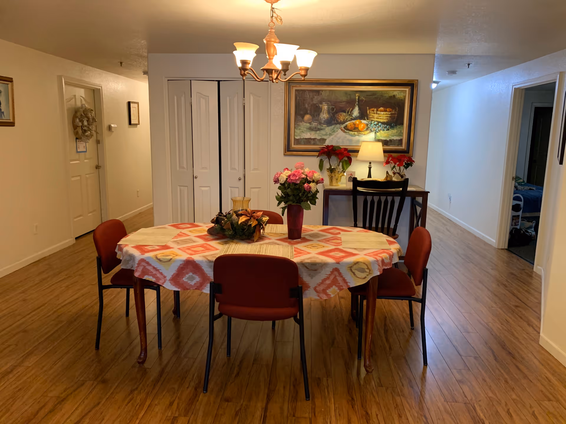 A dining area with a round table covered with a patterned tablecloth and surrounded by six red chairs. On the table are floral arrangements and a decorative centerpiece. Behind the table is a small desk with a lamp, flowers, and a large framed painting on the wall. The room has wooden flooring and white walls, with doorways and closets visible in the background.