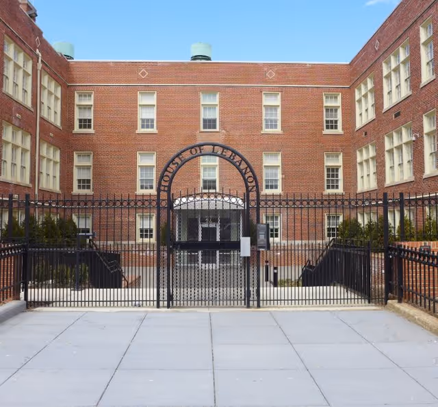 Exterior view of a red brick building with multiple windows and a black metal fence with a gate in front. The gate has an arch with the words 'House of Detention' above it. The sky is clear and blue.