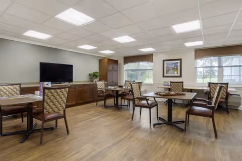 A well-lit common area with several tables and chairs arranged on a wooden floor. There is a large flat-screen TV mounted on a wooden cabinet against the wall, with windows letting in natural light and a framed picture hanging on the wall.