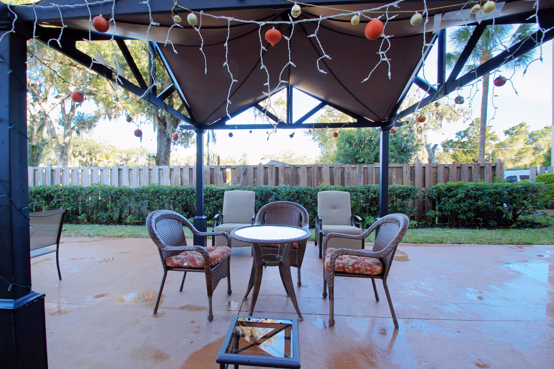 Outdoor patio area with a canopy overhead decorated with hanging string lights and ornaments. There are four chairs arranged around a small round glass table on a wet concrete floor. The patio is surrounded by green bushes and a wooden fence, with trees visible in the background.