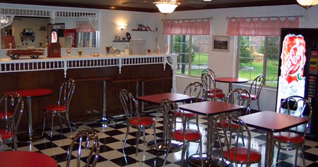 Retro-style dining area with red-topped tables and chrome chairs on a black-and-white checkered floor, a serving counter and a vending machine near windows.