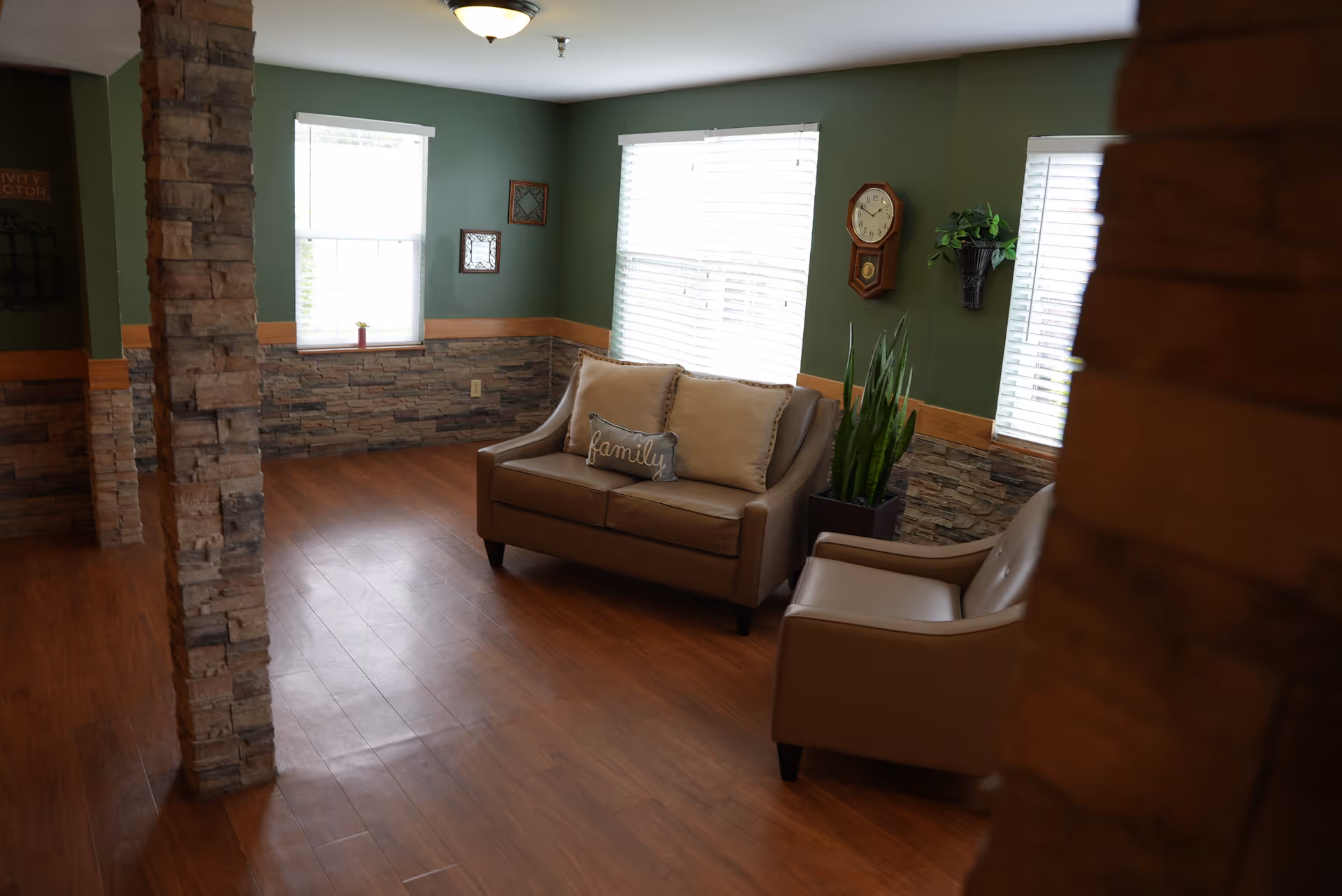 A cozy sitting area with a brown leather loveseat and matching armchair on a wooden floor. The loveseat has two beige pillows, one with the word 'family' embroidered on it. The walls are painted green with a stone accent halfway up, and there are three windows with white blinds letting in natural light. A wall clock and a small plant in a wall-mounted basket are also visible.