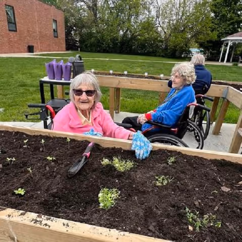 Two elderly women in wheelchairs gardening outdoors at a raised garden bed with small plants and soil. One woman is wearing a pink jacket and blue gardening gloves, smiling at the camera, while the other woman in a blue jacket looks on. There is green grass, trees, and a brick building in the background.