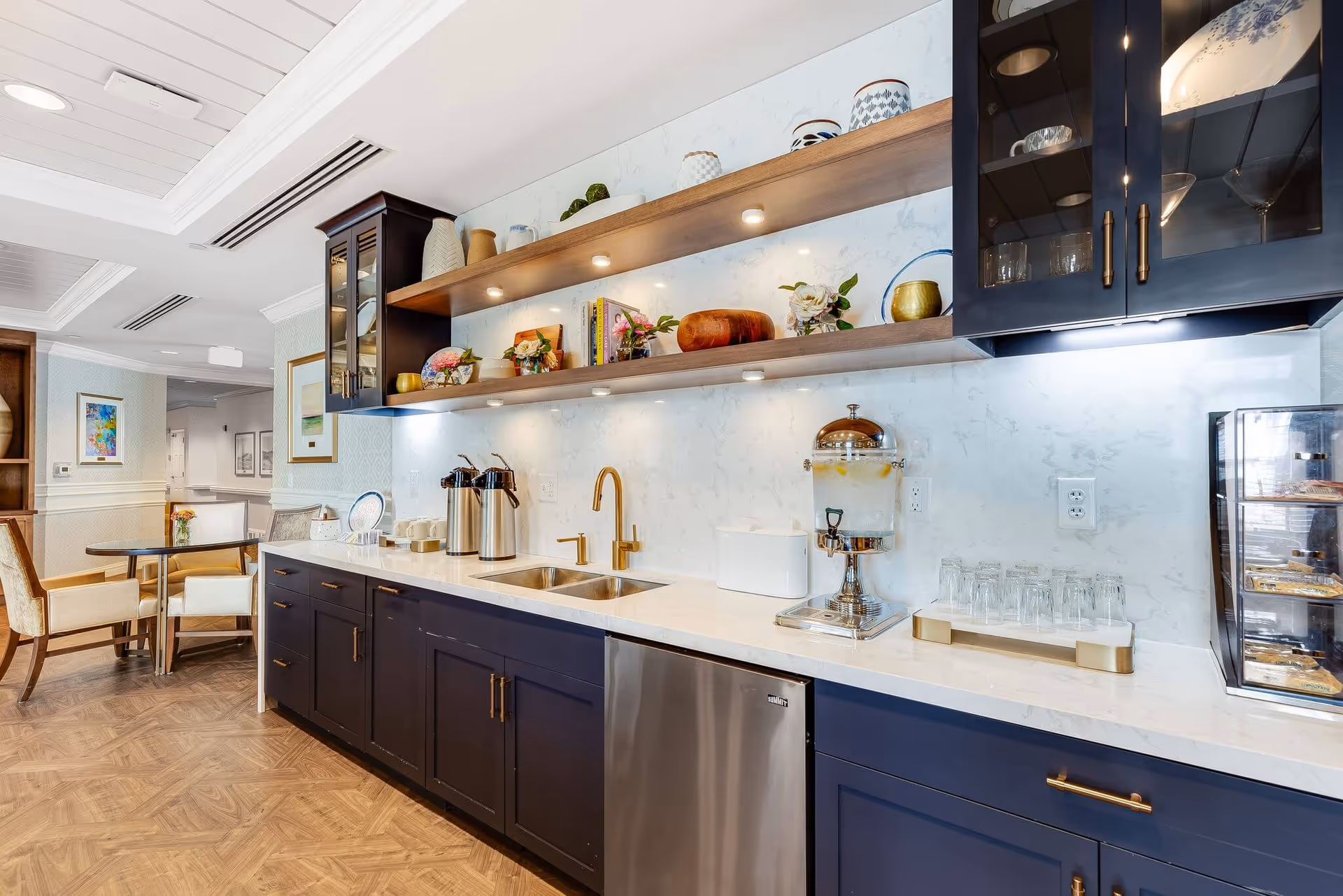 A modern kitchenette/service counter with dark blue lower cabinets, marble countertop, brass faucet, beverage dispensers, open wood shelves with decor, and a small dining area in the background.