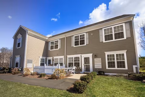 Exterior view of a two-story residential building with beige siding and white trim under a blue sky with some clouds. The building has multiple windows and a small fenced patio area with outdoor furniture. There is a paved walkway and landscaped bushes and grass in the foreground.