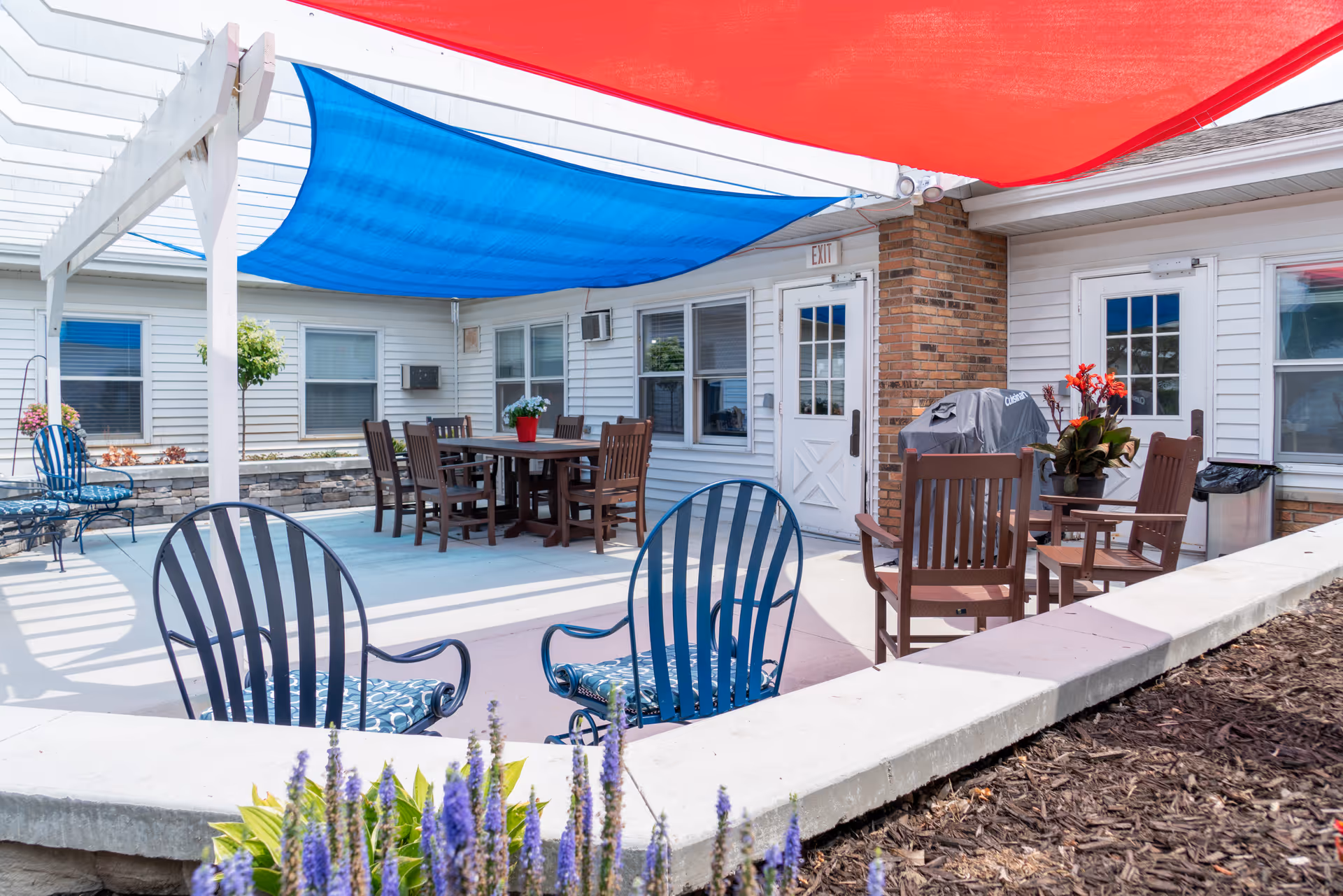 Outdoor patio area with multiple seating arrangements including blue metal chairs and wooden chairs around tables. There are red and blue shade sails overhead, a grill, potted plants, and a white building with windows and doors in the background.