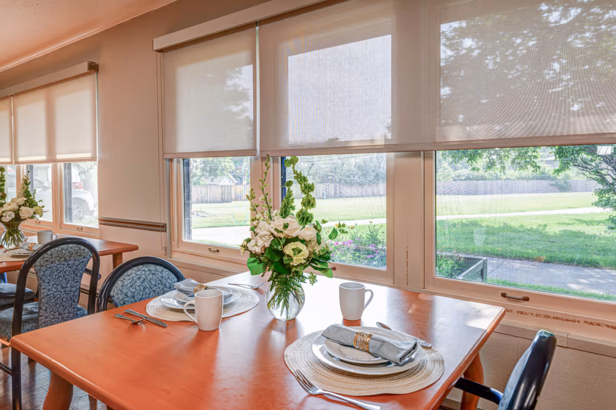 A bright dining area with a wooden table set for two, a vase of flowers, and large windows with blinds overlooking a green lawn.