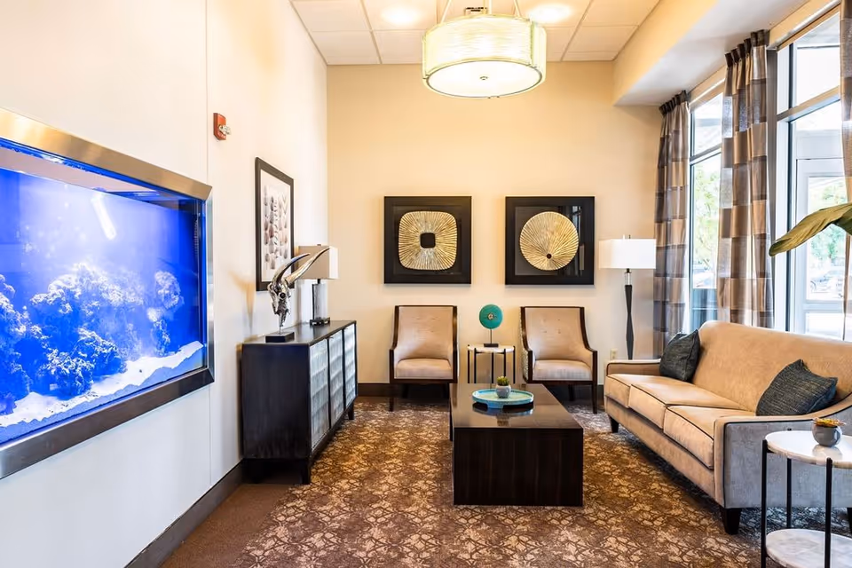 A well-lit living room with a large built-in aquarium on the left wall, a dark wooden cabinet beneath a framed picture, two beige armchairs with a small round table between them, a beige sofa with dark cushions on the right, a dark wooden coffee table with decorative items, patterned carpet, two framed abstract wall art pieces, large windows with checkered curtains, and a ceiling light fixture.