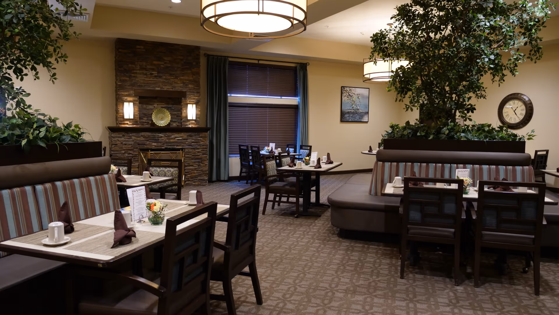 A cozy dining room in a senior living community featuring several tables set with placemats, napkins, cups, and small floral centerpieces. The room has cushioned booth seating with striped upholstery, wooden chairs, a stone fireplace, large windows with blinds, and decorative plants. Warm lighting fixtures hang from the ceiling, and a clock and framed artwork are visible on the walls.