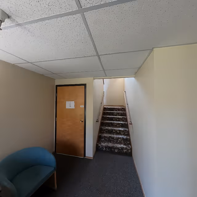 Interior hallway with a carpeted staircase leading upward, a closed wooden door with a sign on it, beige walls, and a blue upholstered chair in the corner.