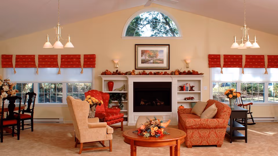 A cozy living room with a vaulted ceiling featuring a large arched window above a white fireplace mantel decorated with autumn-themed garlands. The room has two red armchairs with floral patterns, a beige armchair, and a red patterned sofa with beige cushions. A round wooden coffee table with a floral centerpiece is in the center. On either side of the fireplace are built-in shelves with decorative items. The walls are light beige, and there are large windows with red valances letting in natural light. Two chandeliers hang from the ceiling, and a dining table with chairs is visible on the left side.