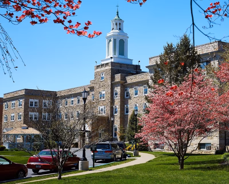 Exterior view of Phoebe Allentown facility showing a large stone building with multiple windows and a white cupola on top. The foreground features a green lawn, a sidewalk, several parked cars, blossoming pink trees, and a clear blue sky.