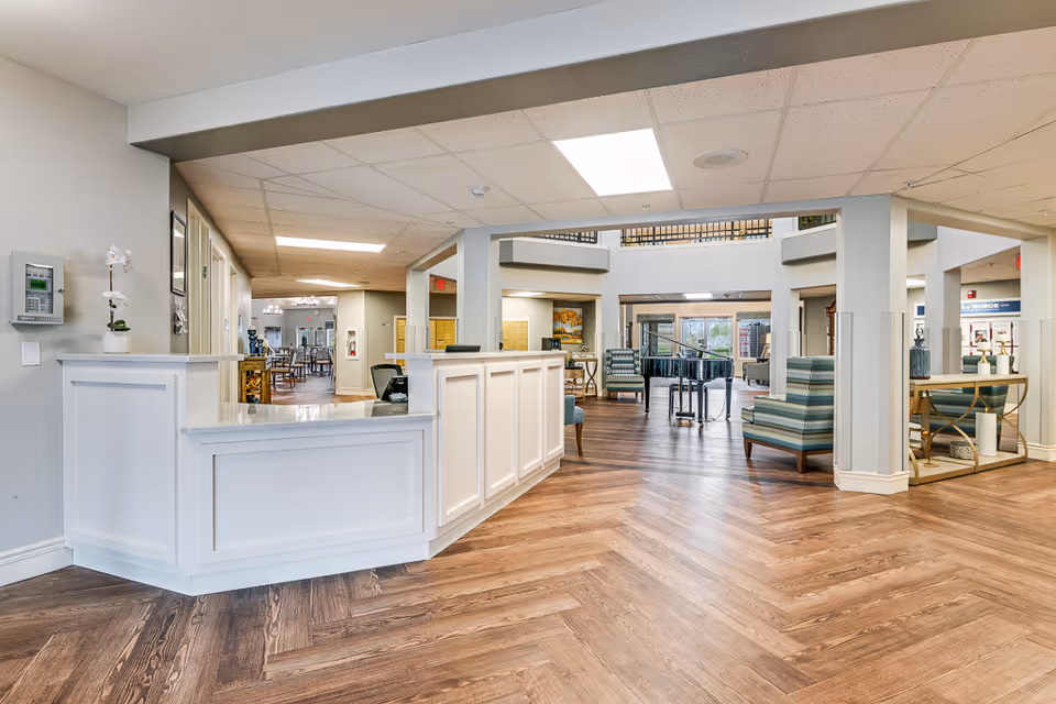 Bright reception lobby with a white check-in desk, seating areas, and wood-patterned floors inside a senior living facility.