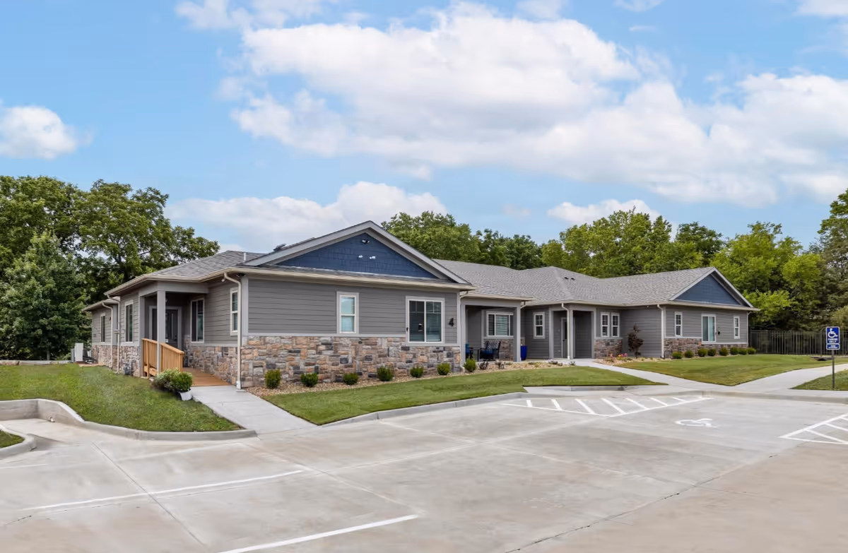 Exterior view of a single-story senior living facility building with gray siding and stone accents, surrounded by green grass and trees under a partly cloudy sky. There is a concrete parking lot in front with marked handicap parking spaces and a sidewalk leading to the entrances.