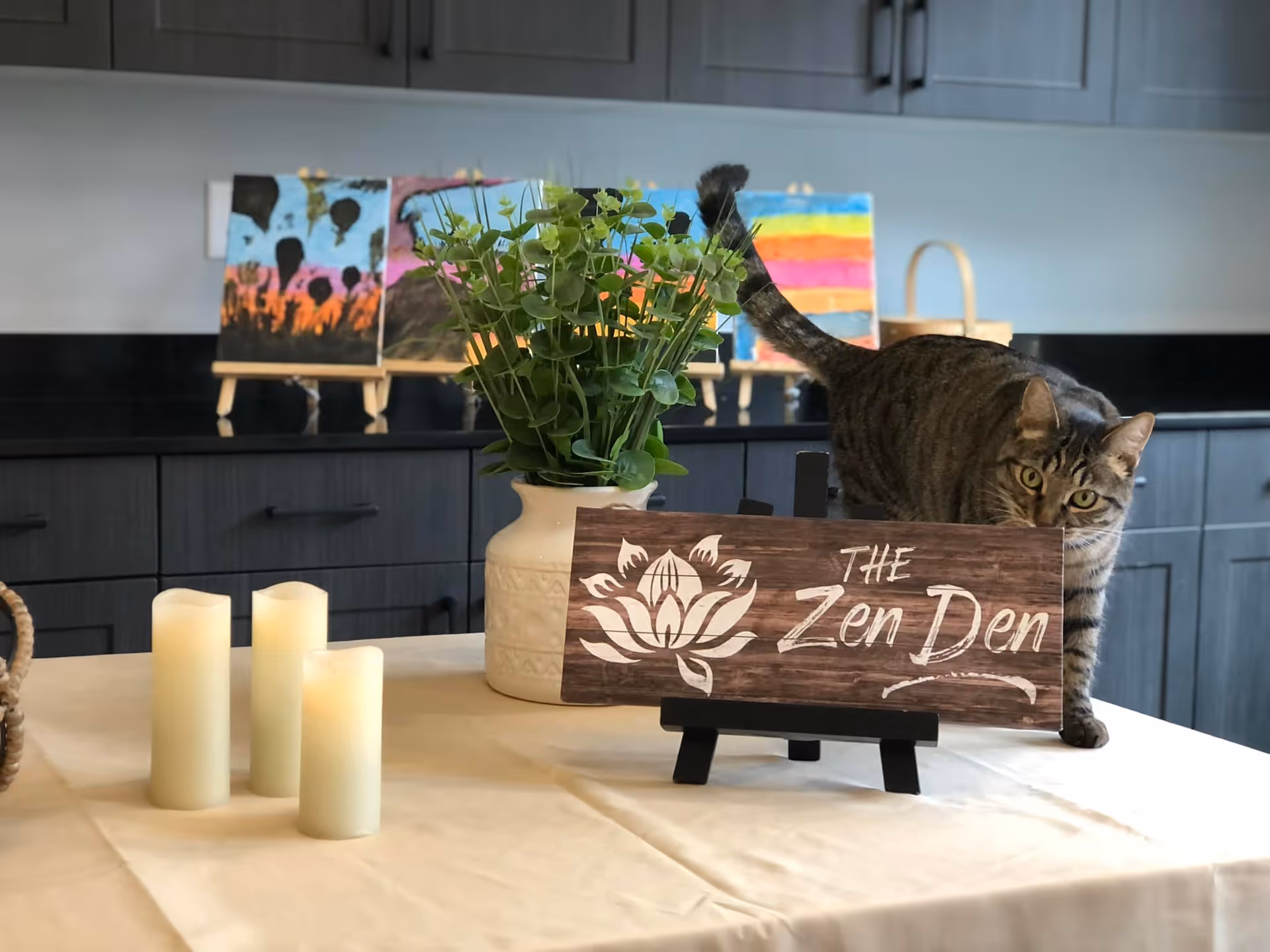 A table with a beige tablecloth displaying four white candles, a white vase with green leafy plants, and a wooden sign that reads 'The Zen Den' with a lotus flower design. A tabby cat is walking behind the sign. In the background, there are colorful paintings on small easels and dark cabinetry.