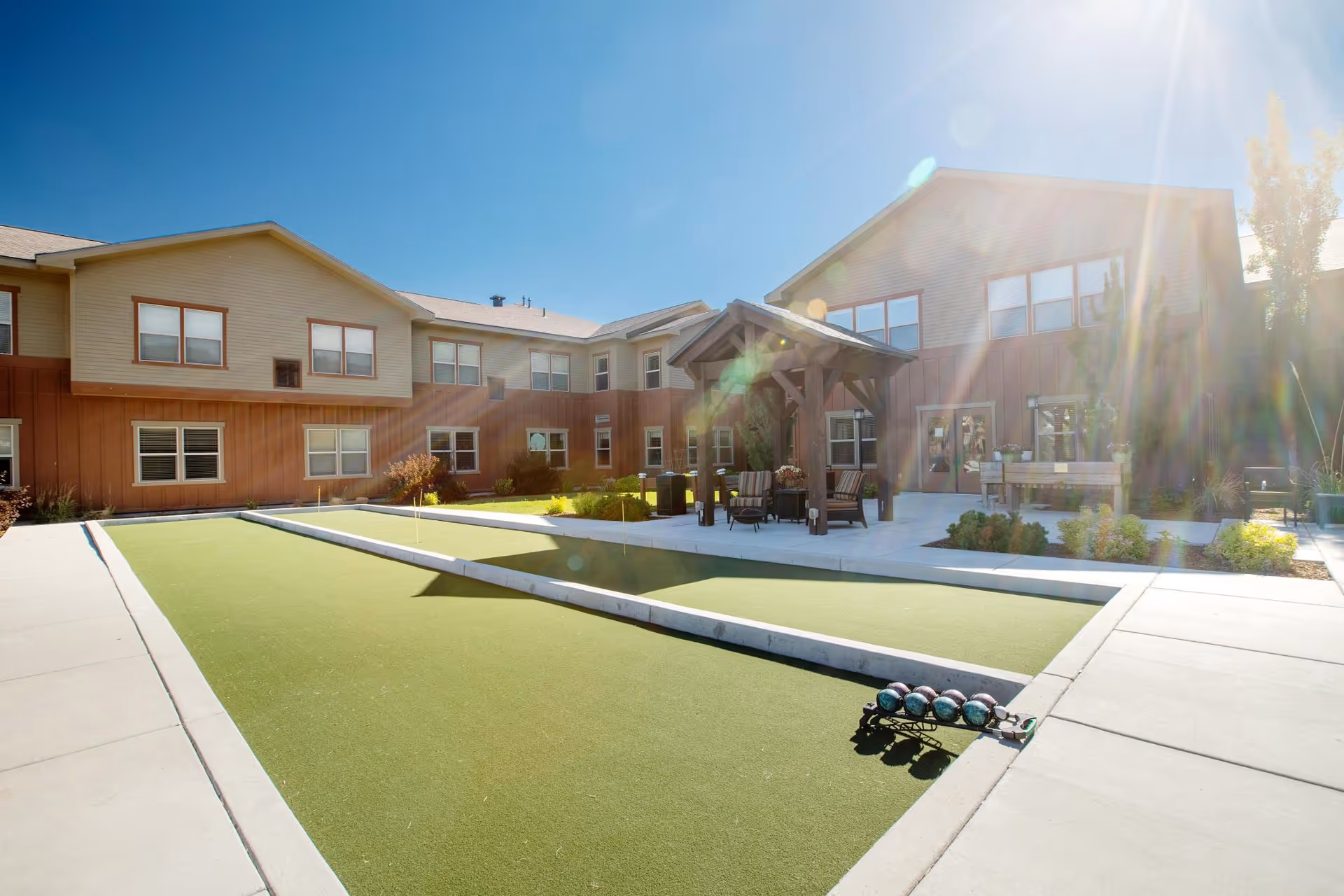 Courtyard with bocce courts, seating under a wooden pergola, and a two-story senior living building in bright sunlight.